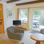 Wohnzimmer mit zwei Sesseln, Tisch, Fernseher und Glaswand zum Garten.