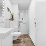 Badezimmer mit Dusche, Waschbecken und Toilette. Weiße Fliesen und Holzfliesen.