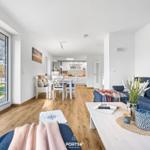 Wohn- und Essbereich mit Holzboden und großem Fenster. Sofa, Tisch und Küchenbereich sichtbar.