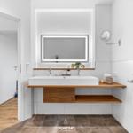 Bad mit Waschbecken, Spiegel und Holzregalen. Fenster und Handtuchhalter sichtbar.