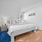 Schlafzimmer mit Bett, Nachttisch und Fenster. Blauer Teppich und Wandbilder.