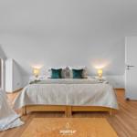 Schlafzimmer mit Bett, Nachttischen und gelbem Sofa. Holzboden und große Fenster.