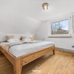 Schlafzimmer mit Holzbett, Nachttisch und Sessel. Fenster mit Blick auf Bäume.