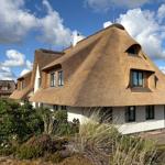 Haus mit strohgedecktem Dach und weißen Wänden, umgeben von Grünflächen und Blumen.