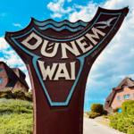 Willkommensschild für Dunenweg mit traditionellen Häusern im Hintergrund.