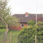 Rotes Backsteingebäude mit Dach und Schornstein hinter Bäumen und Sträuchern.