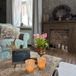 Holztisch mit Tulpen, Kerzen und Schild. Im Hintergrund Sofa und Kamin.