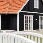 Black wooden house with red roof and white windows. White railing in the foreground.