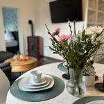 Esstisch mit Blumen, Tassen und Platten. Im Hintergrund Wohnzimmer mit Sofa und Kaminofen.