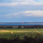 Weitläufige Landschaft mit Leuchtturm, Windkraftanlagen und grünen Feldern am Wasser.