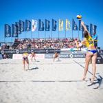 Beachvolleyball-Spiel mit Zuschauern und Sponsorenflaggen am Strand.