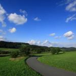 Eine asphaltierte Straße schlängelt sich durch grüne Wiesen unter einem blauen Himmel mit weißen Wolken.