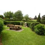 Garten mit Holztischen, Rasenfläche und Hecken. Im Hintergrund sind Bäume und Gebäude sichtbar.