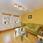 Wohnzimmer mit grünem Sofa, Glas-Tisch und Holzboden. Zwei Fenster und gelbe Wand.