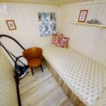 Ein Raum mit Bett, Stuhl und Fenster. Wandregal und Staubsauger sind sichtbar.