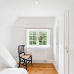 Schlafzimmer mit Dachschrägen, weißer Wand, Holzboden und kleiner schwarzer Stuhl vor Fenster.