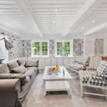 Wohn- und Essbereich mit Sofa, Sesseln und Tisch. Weiße Decke mit Balken. Fenster mit Blick auf Grün.