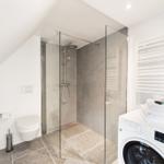 Dusche mit Glaswand, Toilette und Waschmaschine in weißer Umgebung.