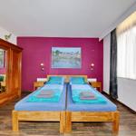 Zwei Betten mit blauen Laken stehen vor einer magentafarbenen Wand. Holzmöbel und Fenster mit Vorhängen.