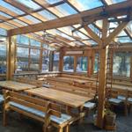 Holzterrasse mit Tischen und Bänken unter Glasdach. Außenbereich mit Blick auf Garten.