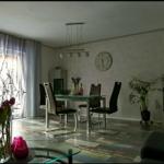 Essbereich mit Tisch, Stühlen und Pflanzen. Fenster mit Vorhängen. Wanduhr und Deckenleuchte.