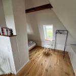 Zimmer mit Holzboden, Bett, Stuhl und Kleiderständer unter Dachschräge.