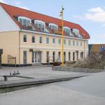 Yellow building with red roof and balconies. Front has shops and parking lot.