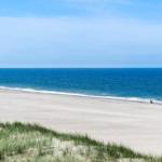Weiterer Blick auf den Strand mit blauem Meer und Sanddünen im Vordergrund.