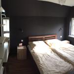 Schlafzimmer mit Bett, Nachttisch und Fenster. Dunkle Wände und Holzboden.