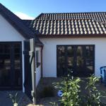 Wohnhaus mit weißen Wänden, dunklem Dach und großzügigen Fenstern. Holzterrasse mit blauer Bank.