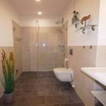 Bad mit Dusche, WC und Waschbecken. Pflanze und Glasregale sind sichtbar.