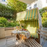 Terrasse mit Strandkorb, Tisch und Stühlen. Hintergrund: grüne Pflanzen.