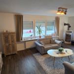 Wohnzimmer mit Sofa, Sessel, Couchtisch und Kaminofen. Holzboden und Fenster mit Jalousien.