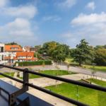 Blick vom Balkon auf Grünflächen, Straßen und Gebäude unter blauem Himmel.