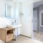 Badezimmer mit Waschtisch und Spiegel. Durchgang zur Dusche.