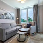 Wohnzimmer mit Sofa, Sessel und Tisch. Fenster mit Blick auf Grün. Wandbild und Deckenleuchte.