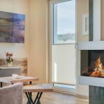 Wohnzimmer mit Kamin, TV und Tisch. Große Fenster mit Jalousien.