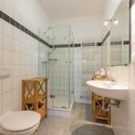 Badezimmer mit Dusche, Waschbecken und Toilette. Weiße Fliesen und Holzregale.