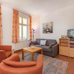 Wohnzimmer mit Sofa, zwei Sesseln, TV und Fenstern mit Vorhängen.