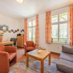 Wohnzimmer mit Sofa, Sesseln, Tisch und Fenstern mit Vorhängen.