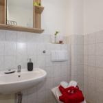 Bad mit Waschbecken, Spiegel und Toilette. Weiße Fliesen und Holzregal.