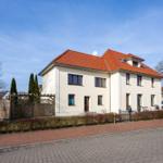 Haus mit rotem Dach und weißen Wänden, Gartenzaun und Straße.