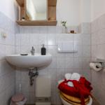 Badezimmer mit Waschbecken, Spiegel und Toilette. Auf dem WC liegt ein roter Bademantel.