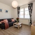 Wohnzimmer mit Sofa, Tisch und Fenster mit floralen Vorhängen.