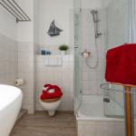 Badezimmer mit Dusche, Toilette und Badewanne. Rote Handtücher und Schiffsmotiv.