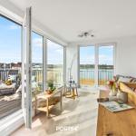 Wohnbereich mit Blick auf Balkon und Wasser. Holztisch, Sofa und große Fenster.