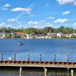 Hafen mit Blick auf einen ruhigen See und eine städtische Uferpromenade mit Häusern und Bäumen.
