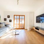 Wohnzimmer mit Sofa, TV und Holzboden. Fenster zur Terrasse.