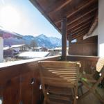 Terrasse mit Holzgeländer und zwei Stühlen. Blick auf verschneite Berge und Dächer.