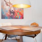 Runder Holztisch mit Stühlen und Platten unter einer Lampe. Wandbild zeigt ein farbenfrohes Reh.